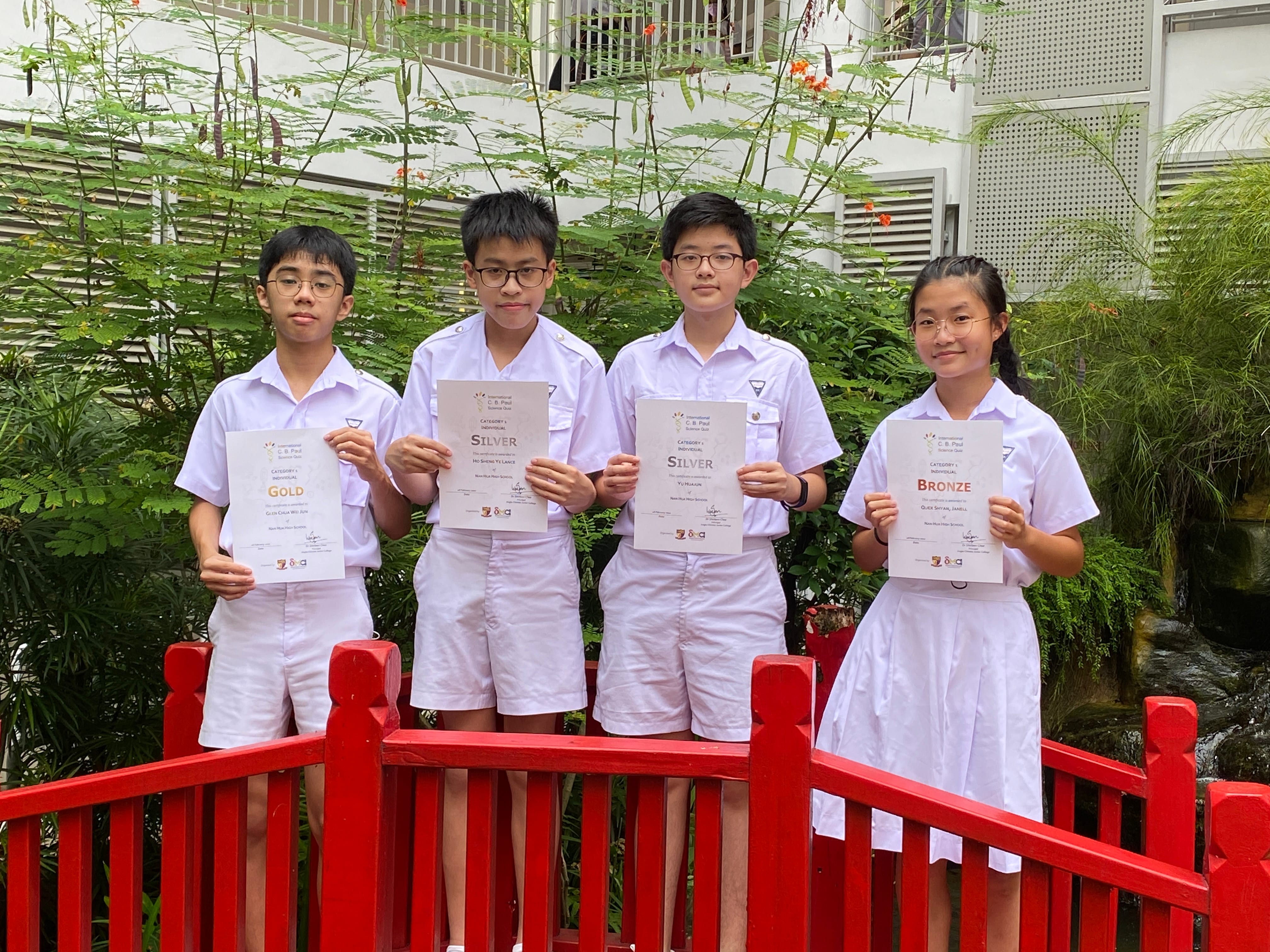 Four students in white uniforms hold Gold, Silver, Silver, and Bronze award certificates on a red bridge.